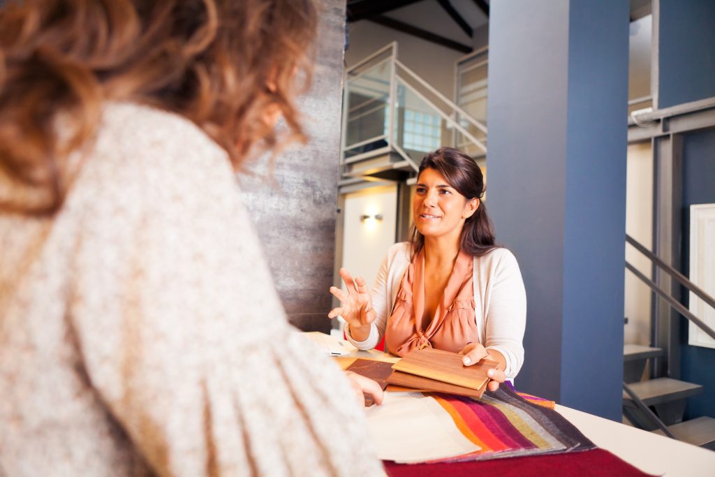 In this image, a showroom consultant is seen showcasing fabric swatches to a customer. The consultant is explaining different fabric options, focusing on custom curtains and blinds that will enhance the customer's home decor. The setting is a stylish showroom, where personalised service is provided to help the customer make informed decisions about her interior design needs. The consultant's engagement with the customer is a tailored approach, offering expertise and suggestions to find the perfect materials that match the customer's preferences and the aesthetics of her home.