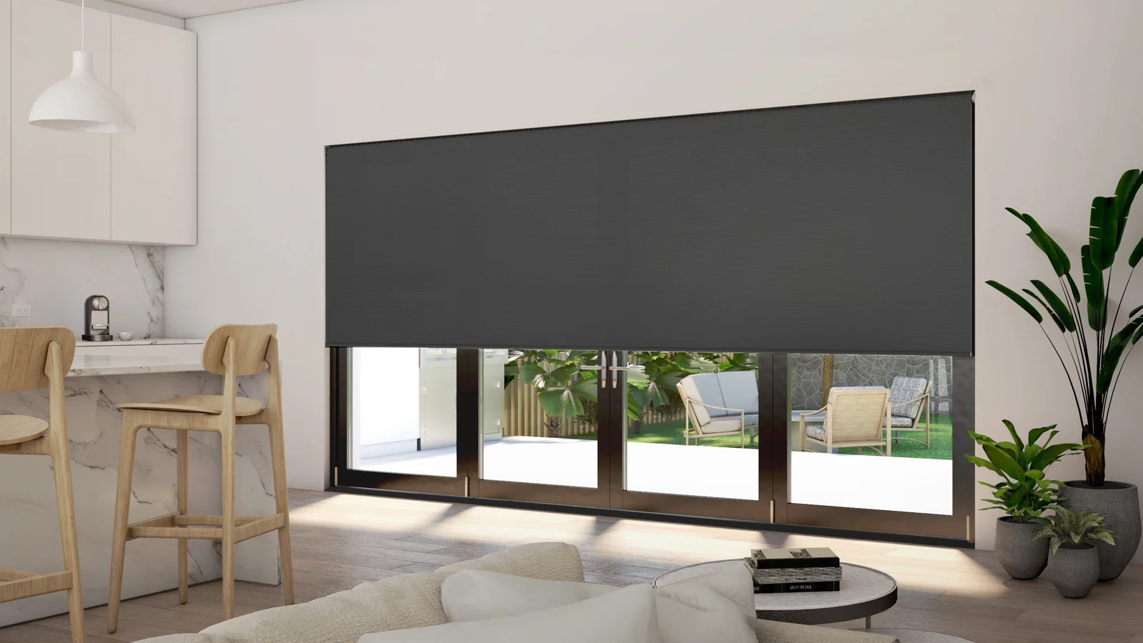 This image features a stylish and modern living space with large windows fitted with sleek black roller blinds that provide a striking contrast to the light and airy interior. The room includes a minimalist kitchen area with white cabinetry and a marble countertop, paired with wooden bar stools for a touch of warmth. A comfortable living area with a plush sofa and a simple coffee table offers a relaxing spot, enhanced by the lush indoor plants adding greenery and vitality to the space. The blinds allow for privacy while still offering glimpses of a tranquil outdoor garden, seamlessly integrating indoor comfort with the beauty of nature outside.