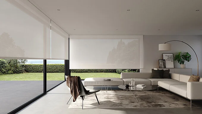 This image shows a luxurious, minimalist living room with large floor-to-ceiling windows covered by sleek white roller blinds. The room features a modern white sectional sofa, a stylish lounge chair draped with a brown throw, and a low coffee table. The interior has a clean aesthetic, emphasized by a polished concrete floor and neutral colors. The windows offer a view of a well-manicured garden, blending indoor and outdoor spaces seamlessly.