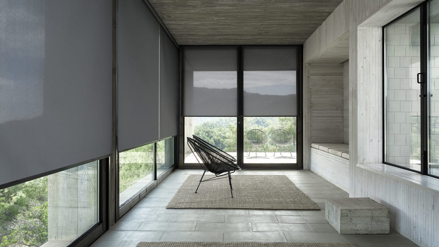 This image showcases a minimalist and contemporary space characterized by large floor-to-ceiling windows fitted with dark gray roller blinds. The room is marked by its raw concrete ceiling and walls, and stone flooring, creating a cool, industrial feel. A unique black wire-frame lounge chair is the central piece of furniture, accompanied by a simple woven rug that adds texture to the space. Outside the windows, a balcony with two circular wire chairs offers a place to enjoy the expansive mountain views. The overall aesthetic is stark yet inviting, emphasizing form and function in a serene, natural setting